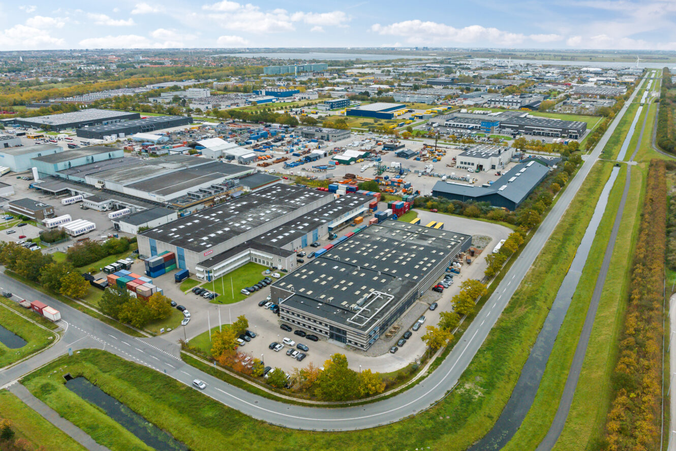 Luftfoto af et industriområde med store lagerbygninger, parkerede biler, skibscontainere og omkringliggende veje, afgrænset af en kanal og grønne områder.