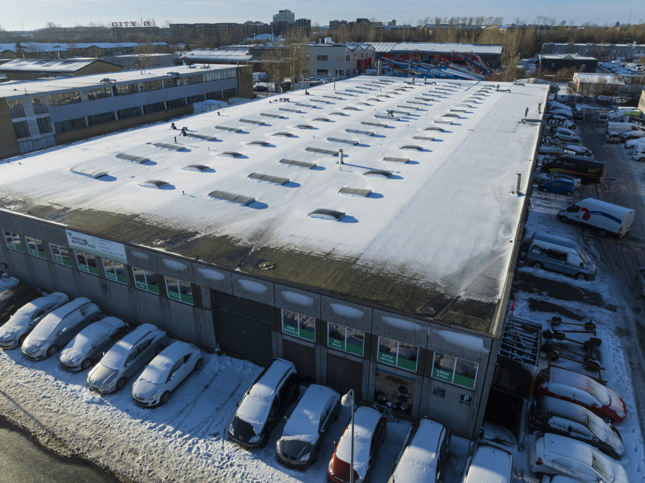 Luftfoto af et stort industrilager med et snedækket tag og parkerede biler, der også er dækket af sne, på en omkringliggende grund.