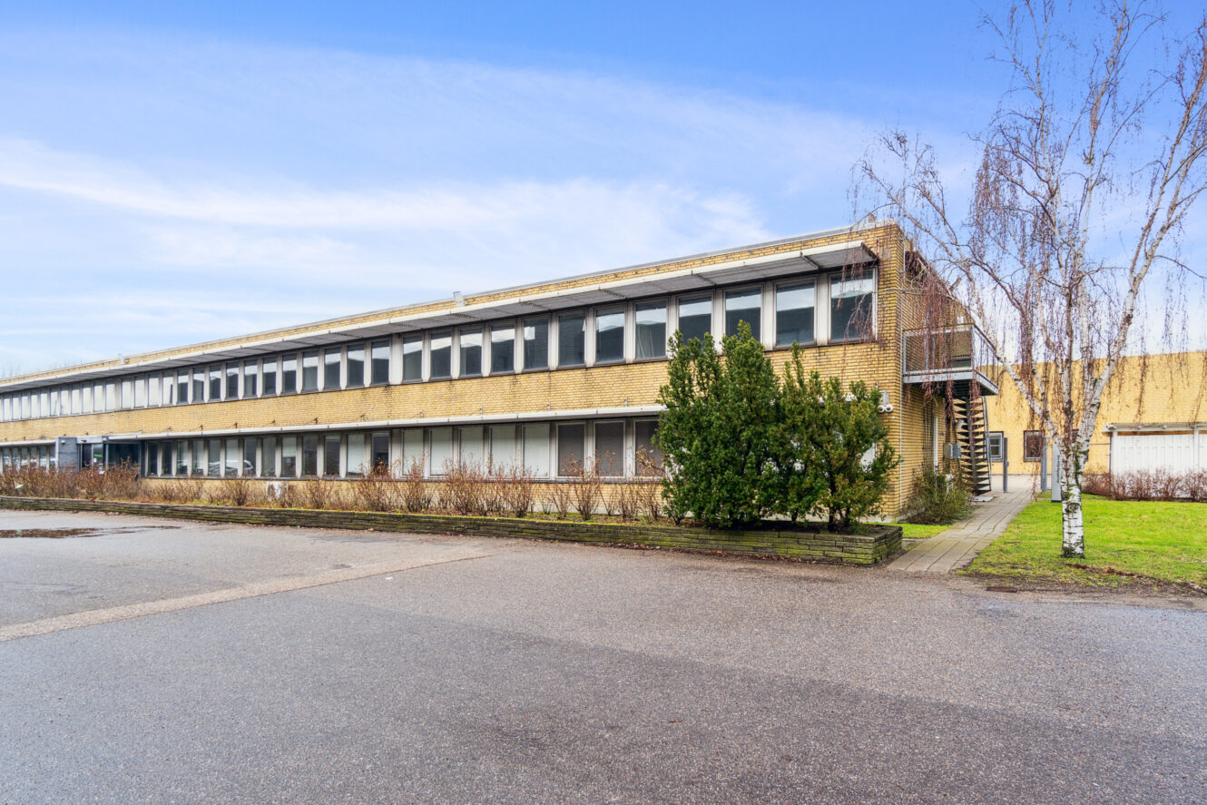 En rektangulær murstensbygning i to etager med mange vinduer, omgivet af buske og træer, under en blå himmel.