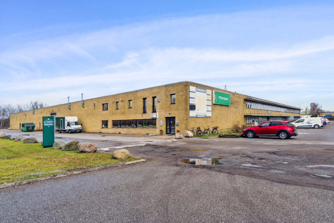 En stor, rektangulær forretningsbygning i gule mursten med flere parkerede biler og cykler foran mod en blå himmel.