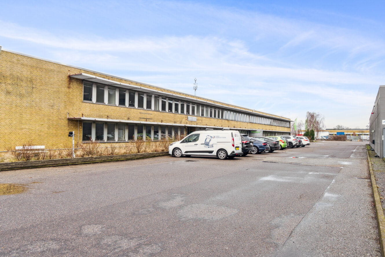 En toetagers kontorbygning i gule mursten med flere parkerede biler foran på en asfalteret plads under en delvis overskyet himmel.