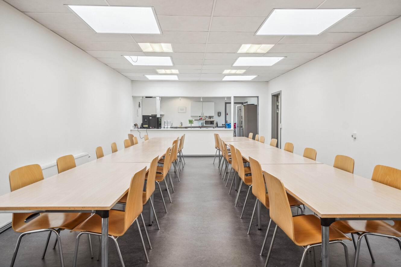Et rent, tomt mødelokale med to rækker træstole og borde, der vender ud mod et køkkenområde bagved. Lyse loftslamper oplyser rummet.