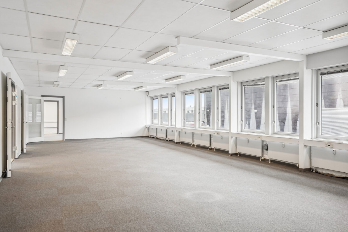 Tomt kontorlokale med store vinduer, hvide vægge, loftslamper og beige gulvtæppe. Rummet er godt oplyst af det naturlige lys, der kommer ind gennem vinduerne.