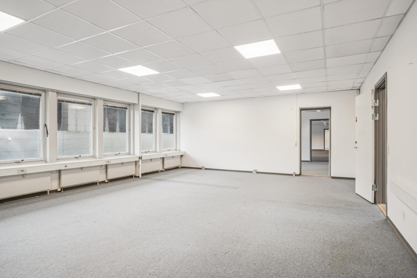 Tomt kontorlokale med gråt gulvtæppe, hvide vægge, store vinduer på den ene side, loftslamper og to døråbninger, der fører til tilstødende lokaler.