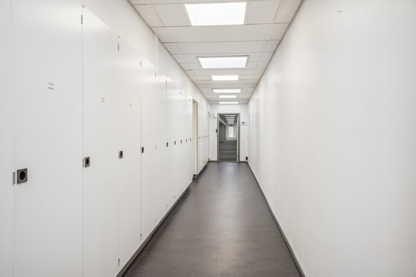 En lang, tom gang med hvide vægge, loftslamper og mørkt gulv, med lukkede opbevaringsskabe til venstre og en trappe for enden.