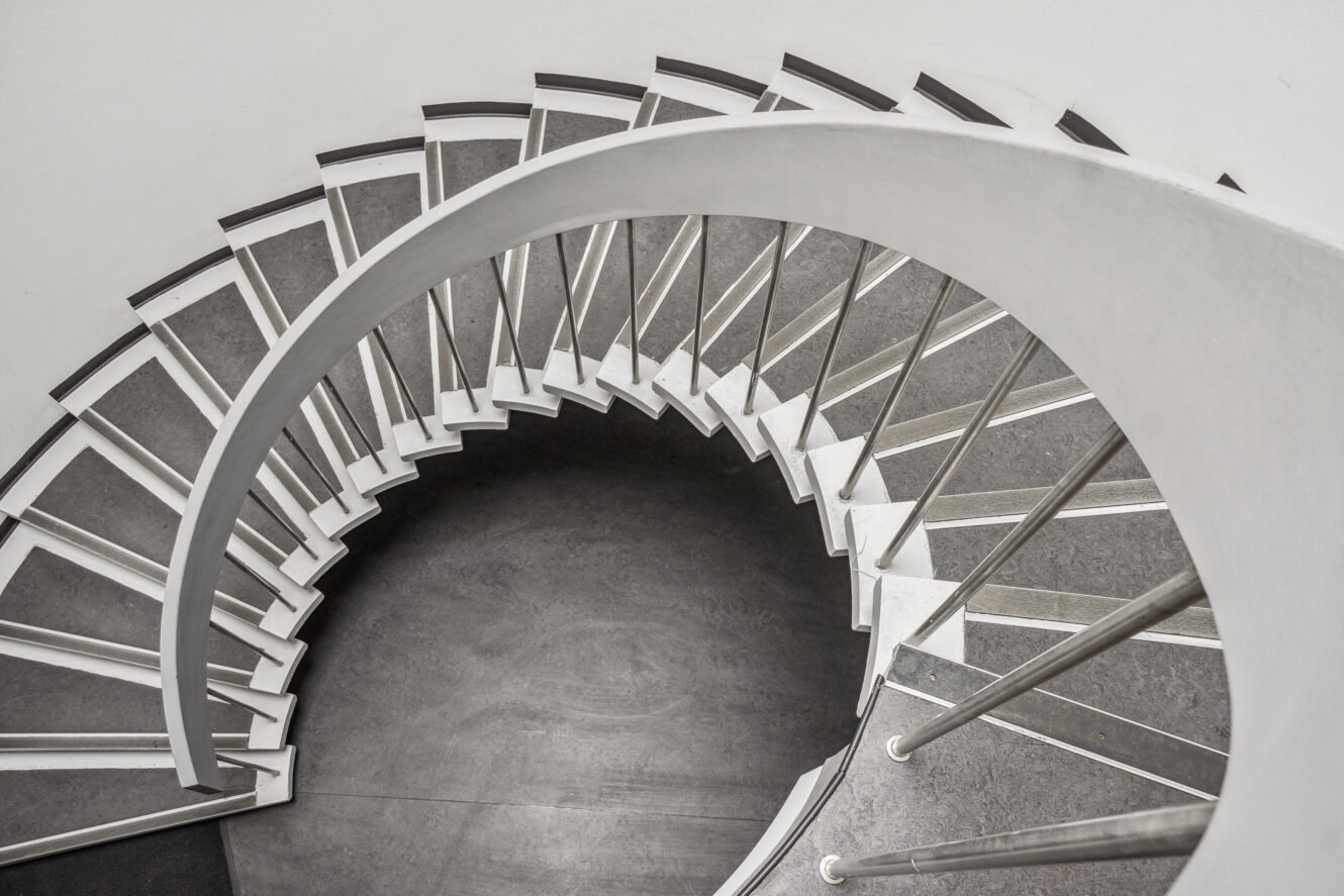 En top-down-visning af en moderne vindeltrappe med mørke trin, hvide søjler og metalgelænder mod en almindelig hvid væg.