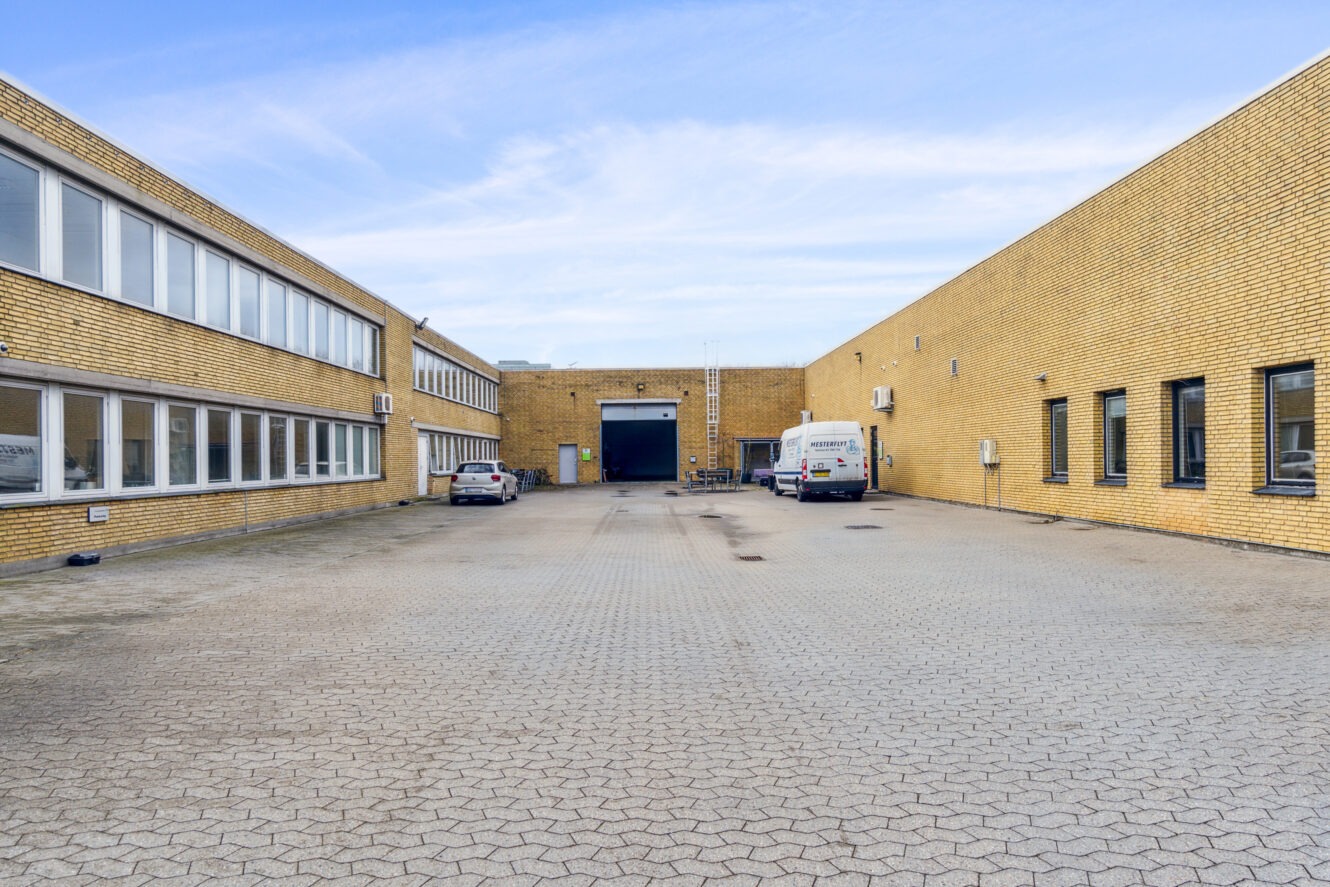 En brolagt gårdsplads mellem to industribygninger i gule mursten med nogle få parkerede biler og en central læsserampe under en delvis overskyet himmel.