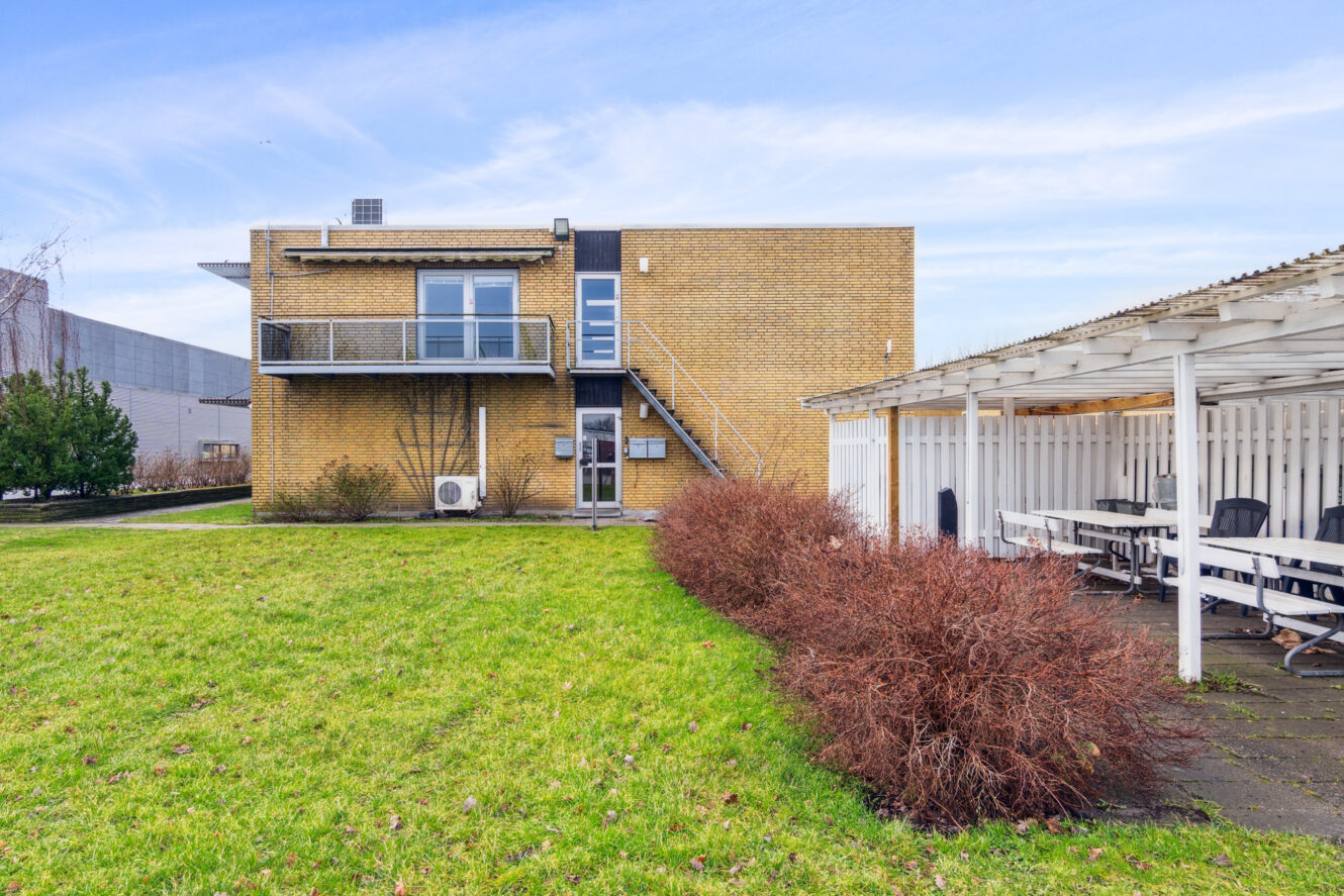 En toetagers gul murstensbygning med udvendige trapper og en balkon, der støder op til et overdækket udendørs opholdsområde på en græsplæne.