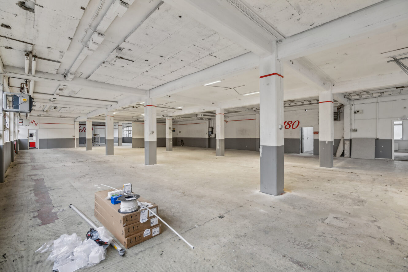 Rummeligt, tomt industrielt lagerinteriør med betonsøjler, grå og hvide vægge, højt til loftet og nogle elektriske forsyninger på gulvet i forgrunden.