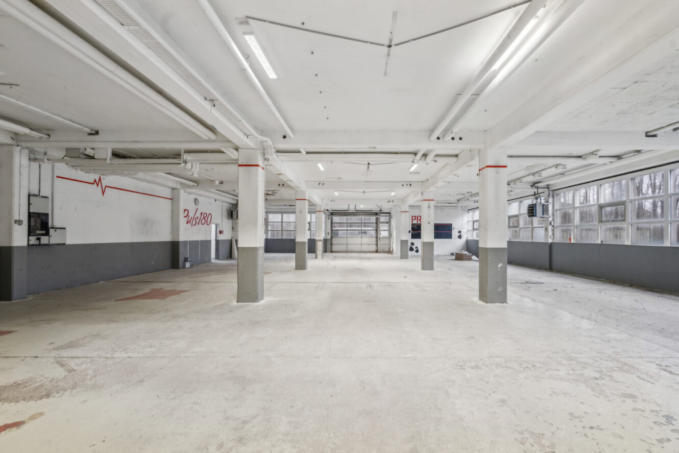 Rummelig, tom industrigarage med hvide vægge, synlige rør, store vinduer og betongulv. På nogle af væggene er der malede linjer og tekst.