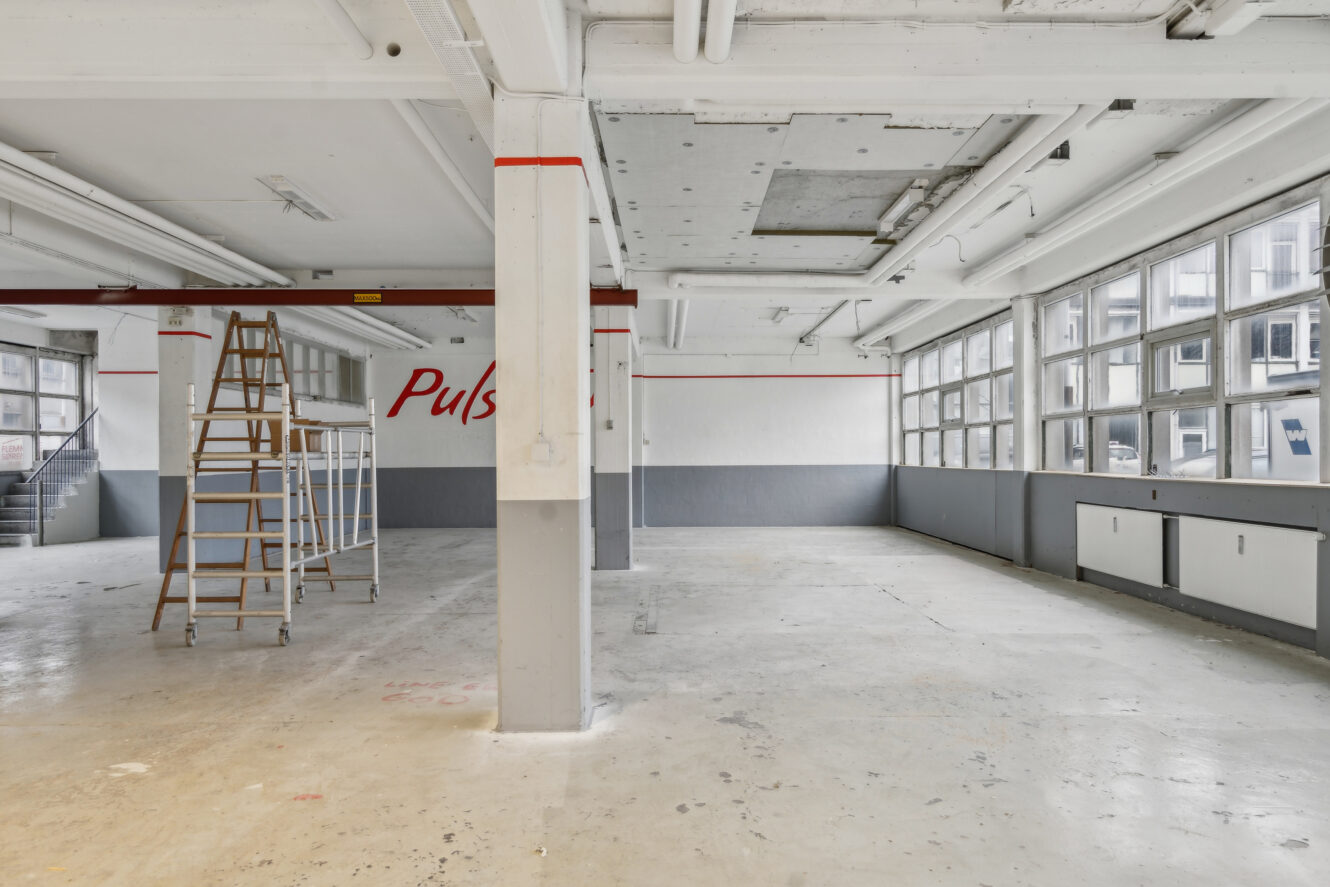Et tomt industrilokale med store vinduer, synlige rør og en træstige tæt på væggen; ordet Puls er delvist synligt på bagvæggen.
