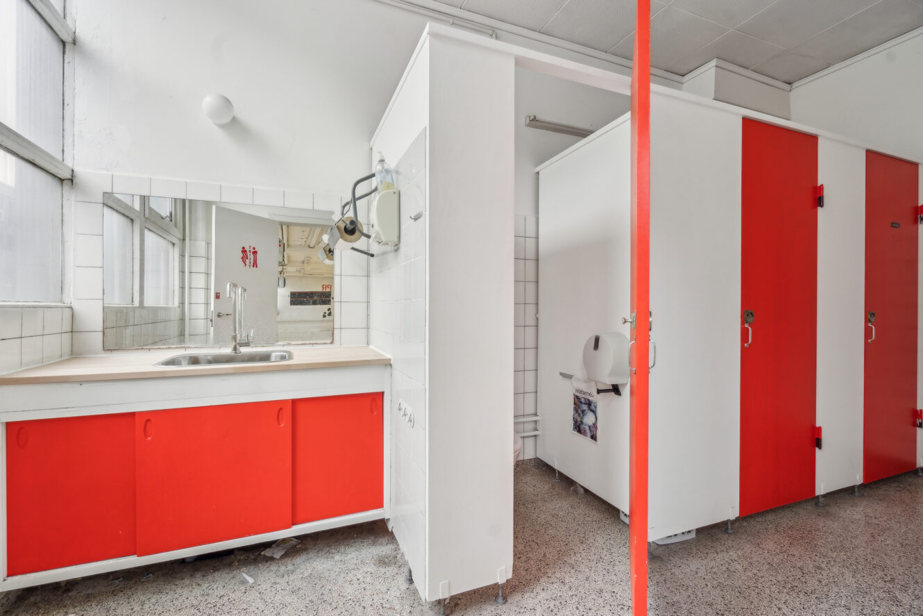 Et offentligt toilet med røde og hvide båse, et stort spejl over vasken og flisebelagte vægge og gulv.