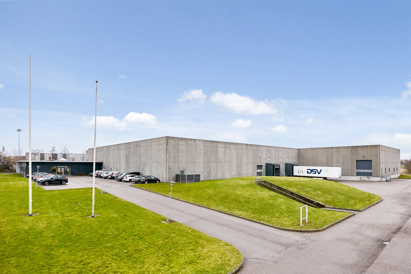 Et stort, moderne industrilager med et ydre af beton er omgivet af grønne plæner, en parkeringsplads med biler og en DSV-lastbil parkeret ved en læsserampe.