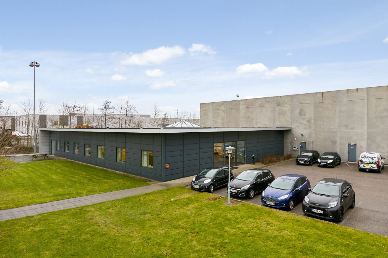 En moderne industribygning i én etage med fladt tag, flere parkerede biler og en græsplæne under en delvis overskyet himmel.