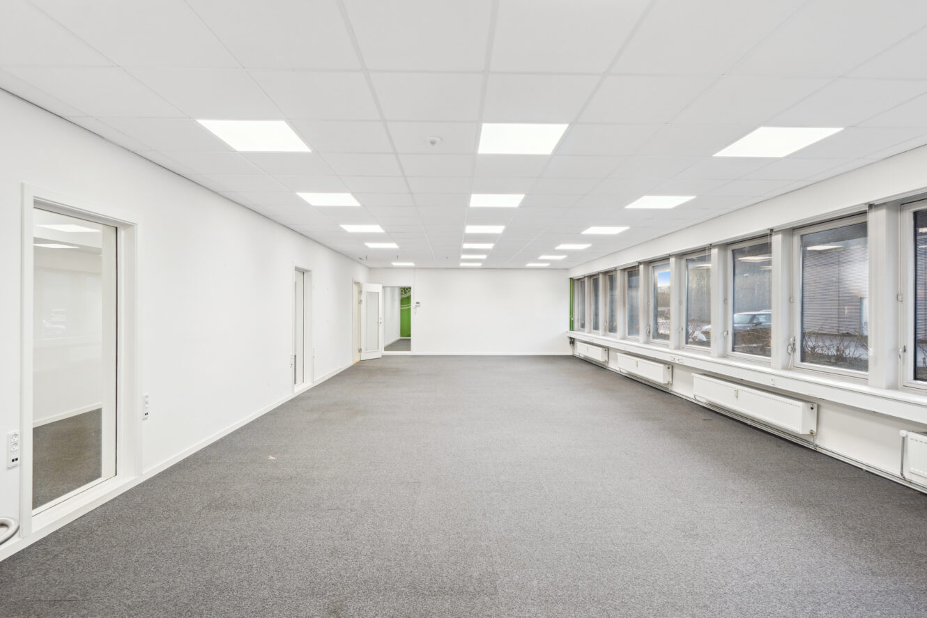 Tomt, moderne kontorlokale med store vinduer på den ene side, hvide vægge, gråt gulvtæppe og loftslamper.