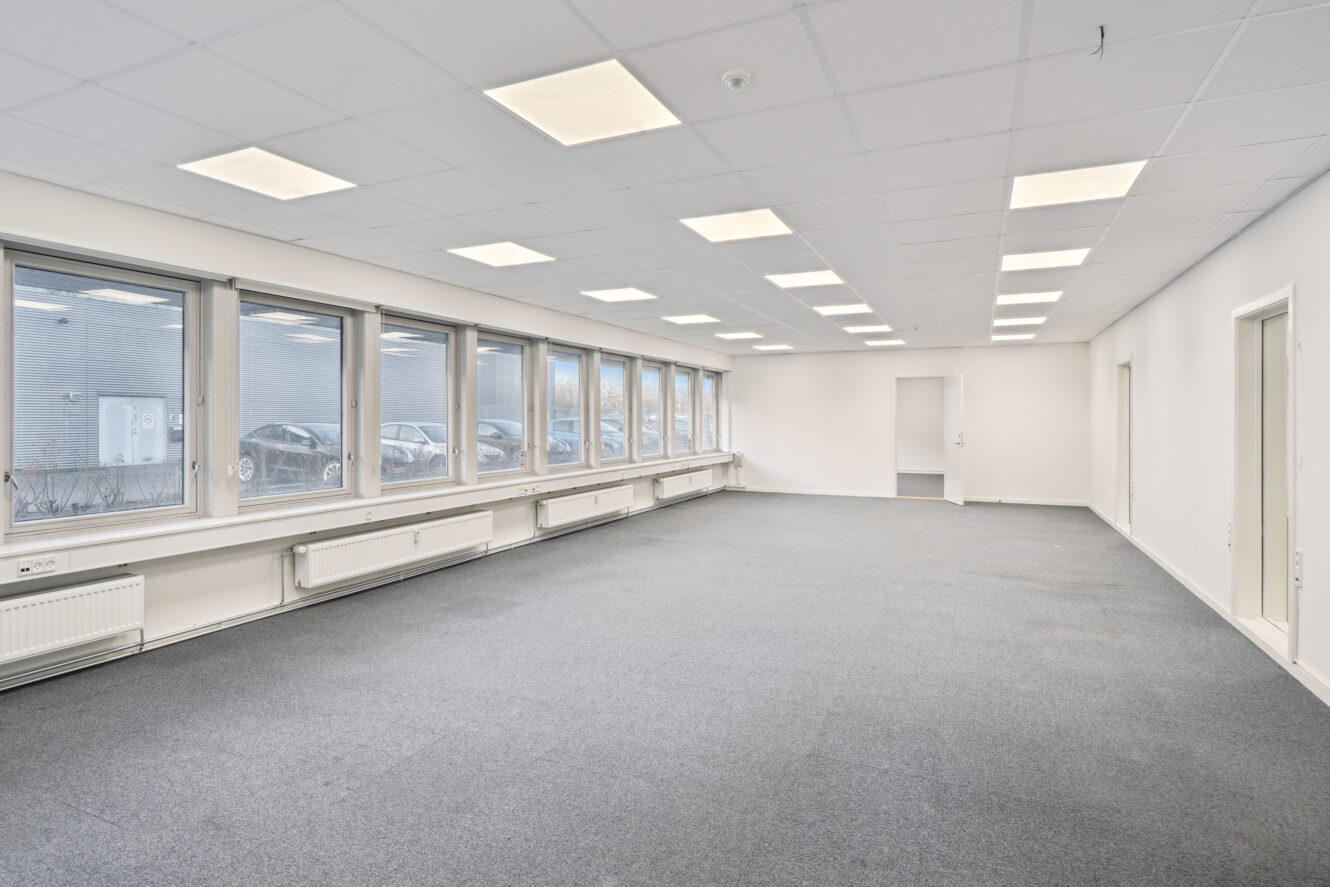 Tomt kontorlokale med gråt gulvtæppe, hvide vægge, store vinduer langs den ene side og loftslamper jævnt fordelt over loftet.