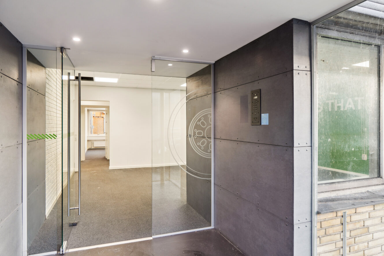 Indgangsdøre af glas fører ind i et moderne kontorlokale med gråt gulv, hvide vægge og kraftig ovenlys. Et samtaleanlæg er monteret på væggen ved siden af døren.