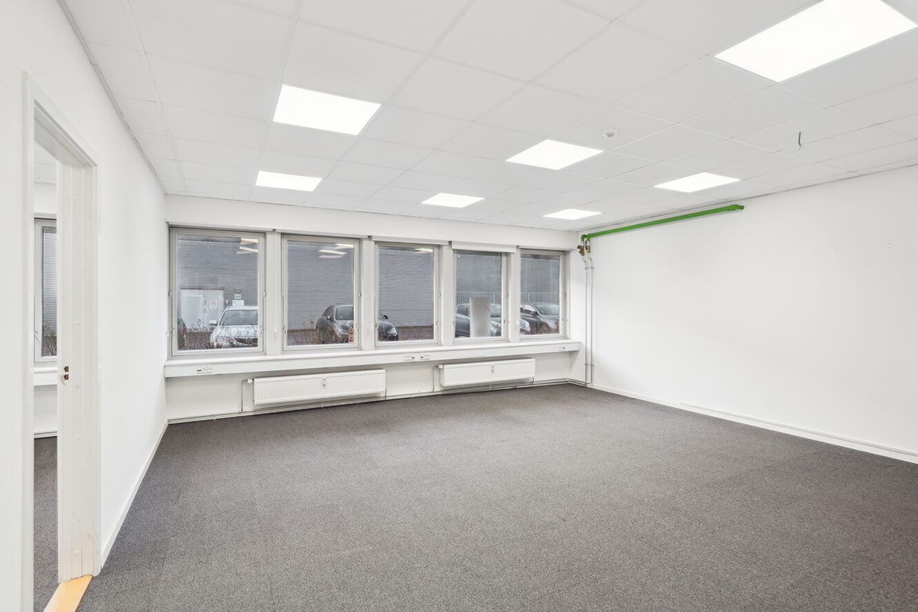 Tomt kontorlokale med hvide vægge, gråt gulvtæppe, loftslamper, flere vinduer, varmeapparater under vinduerne og et grønt rør på den ene væg.