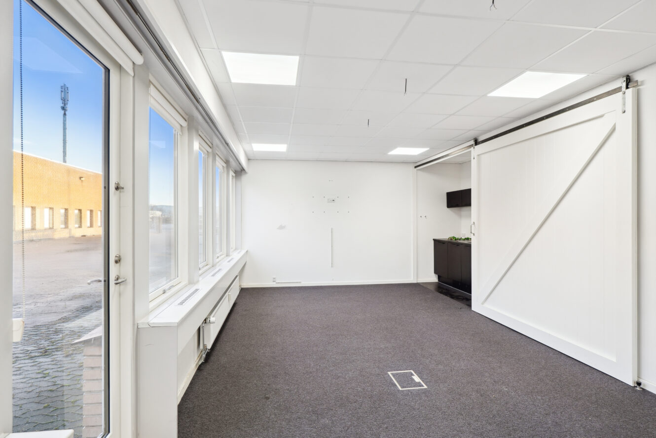 Lyst, tomt kontorlokale med store vinduer, gråt gulvtæppe, hvide loftsplader, en skydedør og et lille, mørkt tekøkken.