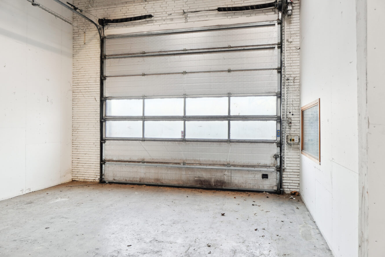 En tom industrigarage med en stor rulleport, hvide murstensvægge, et lille vindue i væggen og et betongulv.
