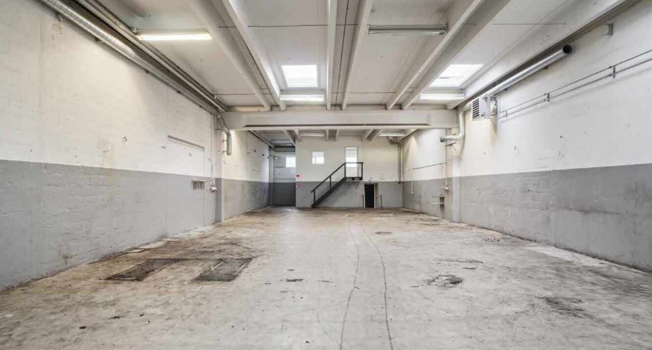 Tomt industrilager med hvide og grå vægge, betongulv, højt til loftet med ovenlysvinduer og en metaltrappe, der fører op til et lille øvre område bagerst.