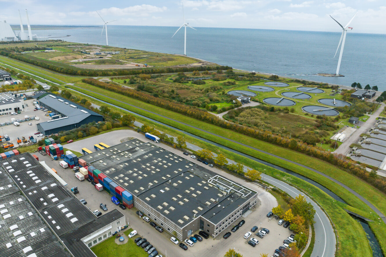 Luftfoto af et industrianlæg nær en kystlinje med vindmøller og cirkulære vandbehandlingstanke i baggrunden; flere lastbiler er parkeret ved siden af lagerbygninger.