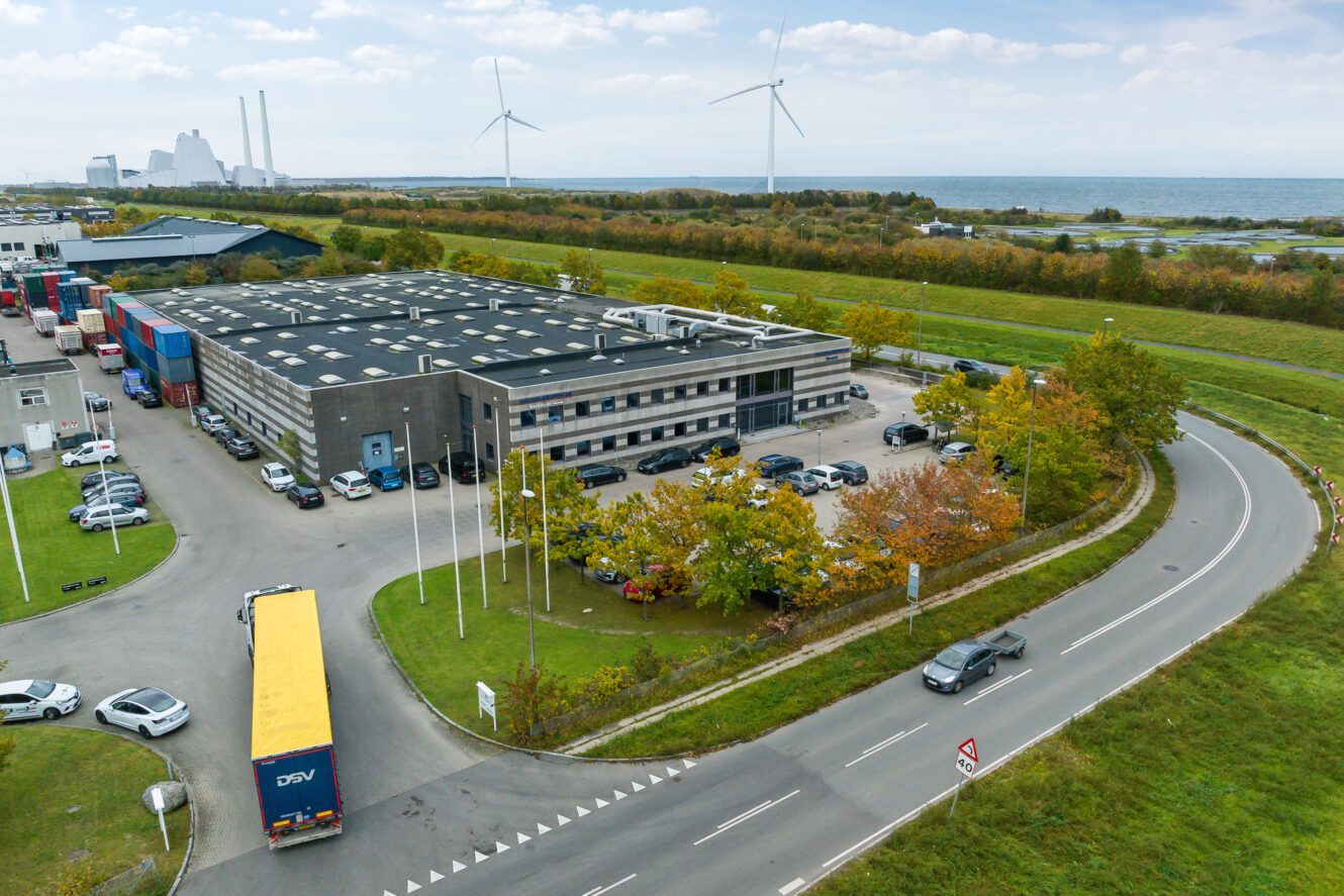 Luftfoto af et industrianlæg med en parkeringsplads, lastbiler, omkringliggende grønne områder, vindmøller og et nærliggende vandområde under en delvis overskyet himmel.
