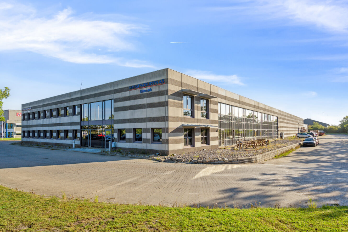 En moderne, toetagers kontorbygning med en stor parkeringsplads og flere biler parkeret udenfor på en solrig dag.