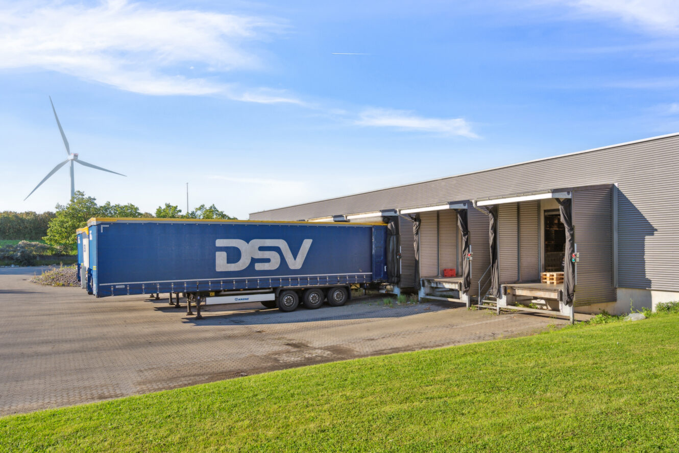 En blå DSV-lastbiltrailer er parkeret ved en læsserampe uden for en moderne lagerbygning, og i baggrunden ses en vindmølle.