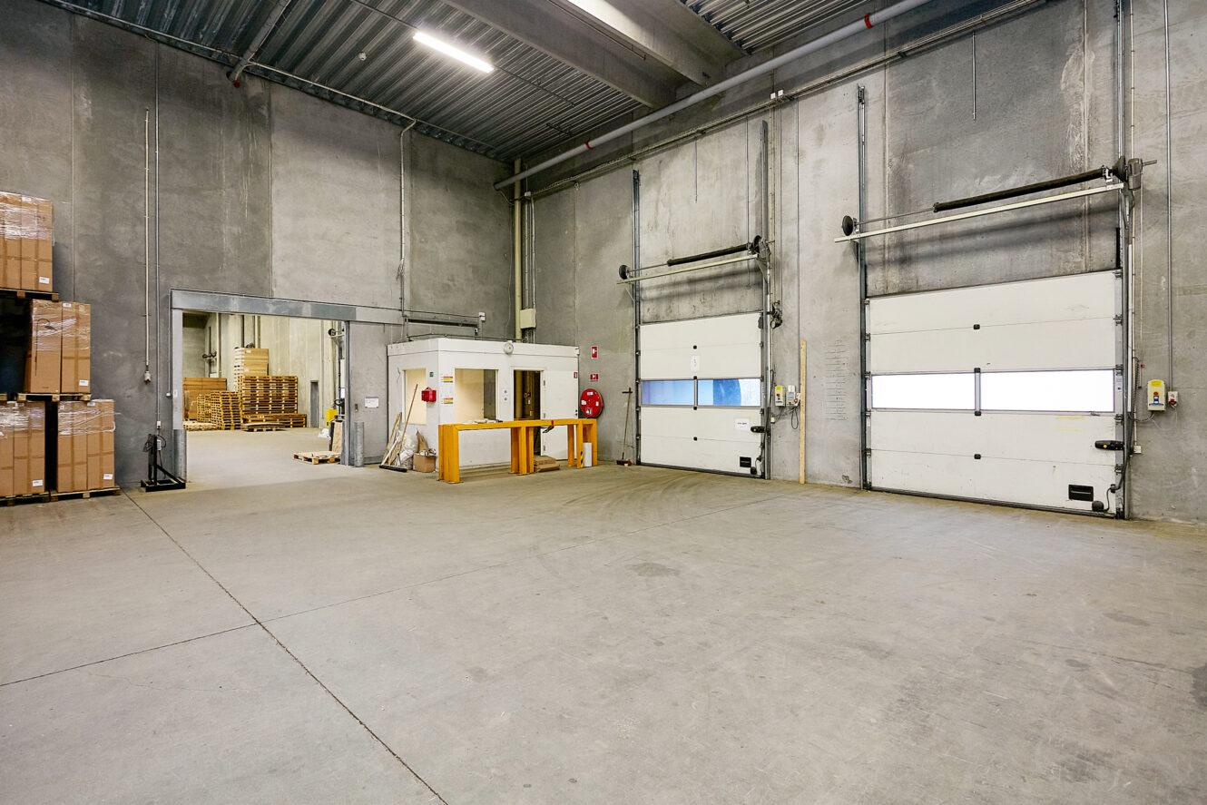 Interiør af en tom lagerhal med betonvægge, tre porte til læsseramper, stabler af paller og kasser og et lille kontorområde bagved.