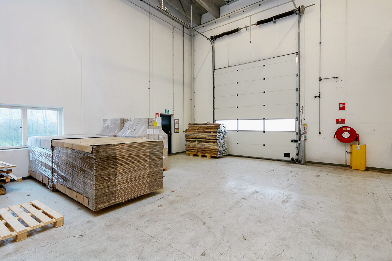 Lageret er indrettet med stablede papark på paller, en stor lukket læsseport, brandsikkerhedsudstyr og et lille vindue, der lukker naturligt lys ind.