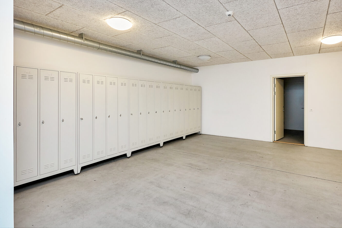 Et rum med hvide skabe langs den ene væg, et betongulv, hvide vægge og en åben dør, der fører ind til et andet rum.