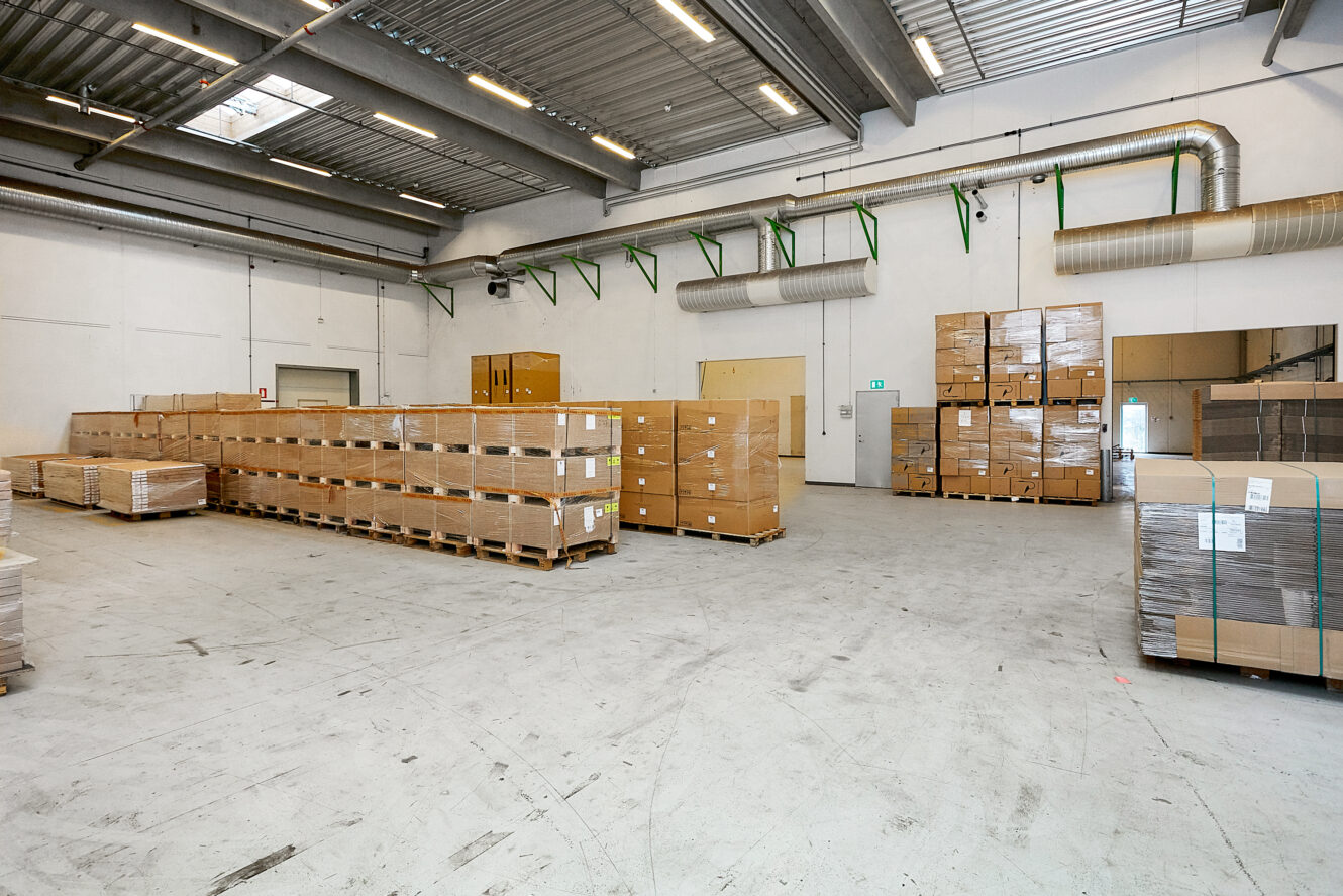 Stort lagerinteriør med stabler af papkasser på træpaller, højt til loftet og fluorescerende belysning.