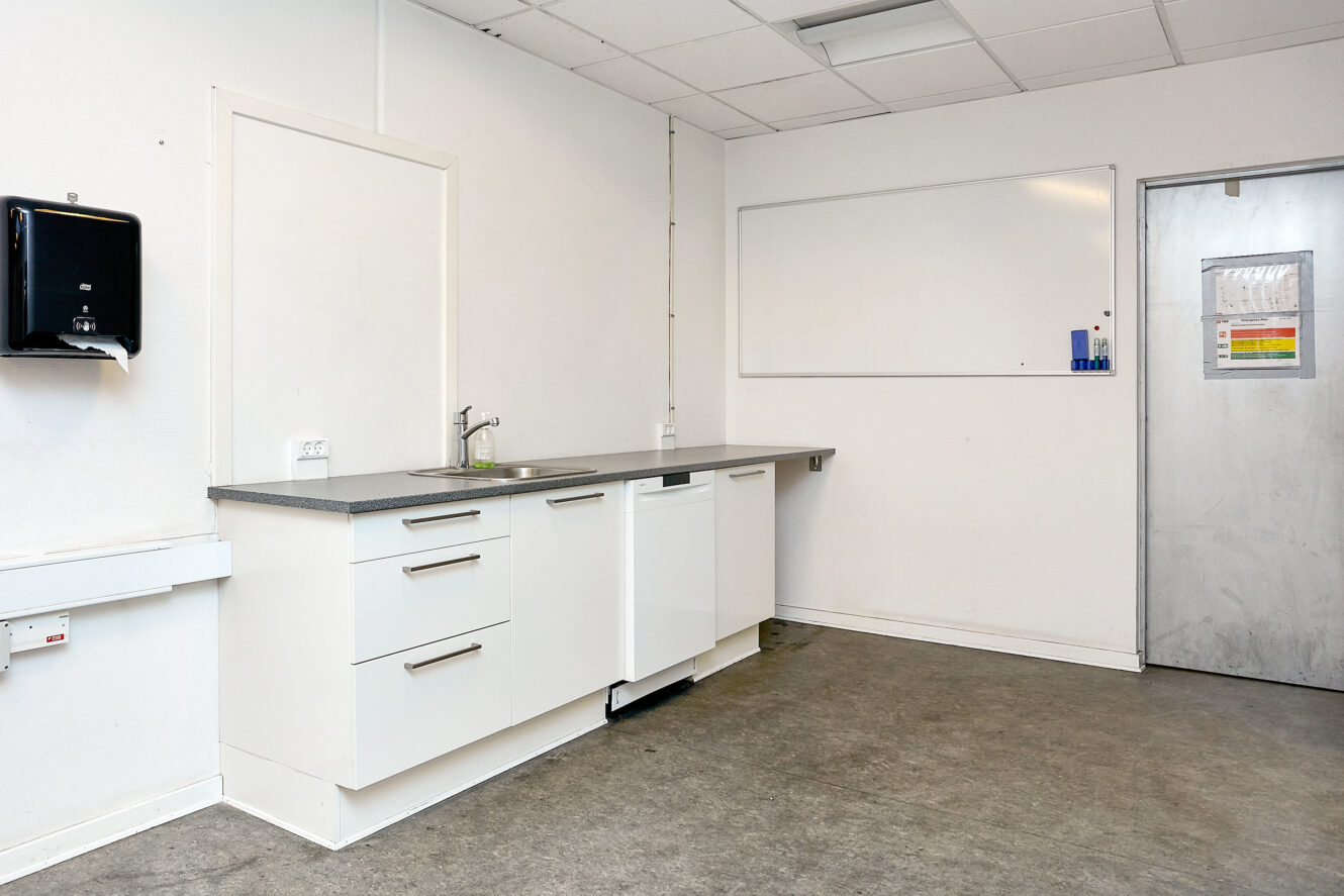 Et enkelt, moderne tekøkken med hvide skabe, vask, opvaskemaskine og whiteboard i et rum med almindelige hvide vægge og et gråt gulv.