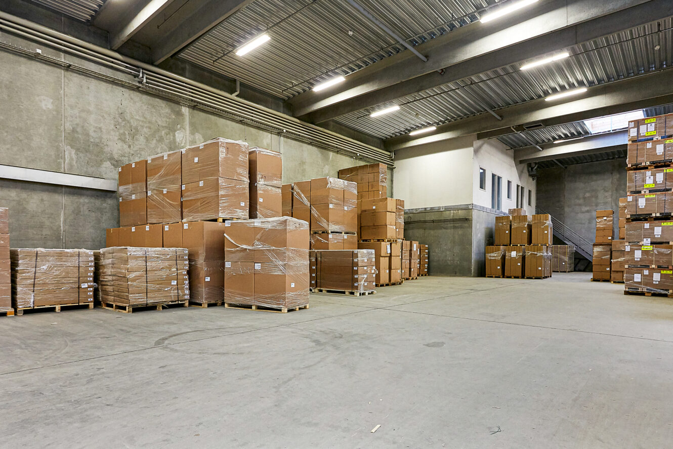 Stort lagerinteriør med stabler af papkasser pakket ind i plastik på paller, arrangeret langs betonvægge under fluorescerende belysning.