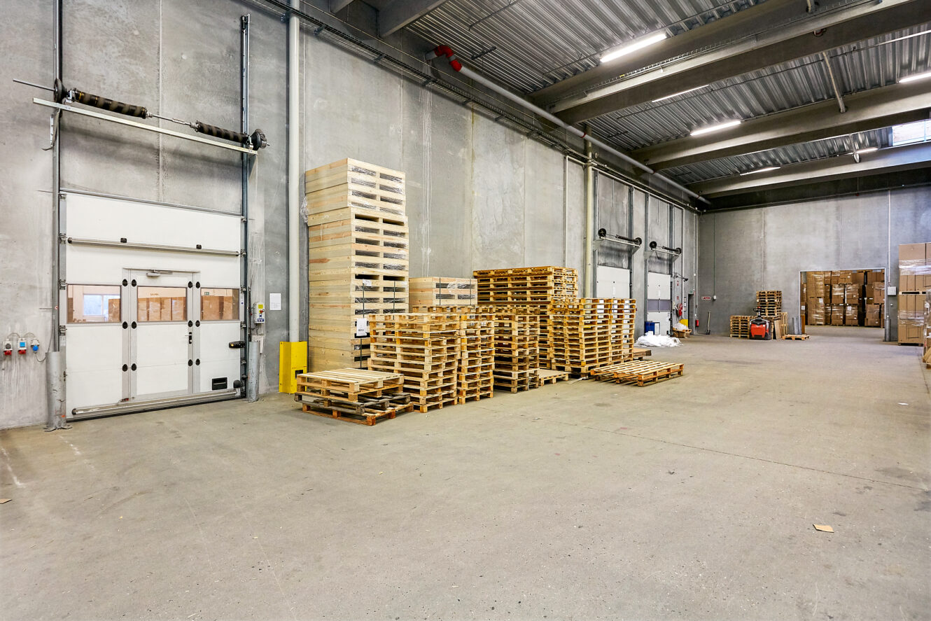 Stabler af træpaller står langs væggen i en rummelig, veloplyst lagerhal med højt til loftet og betongulv.