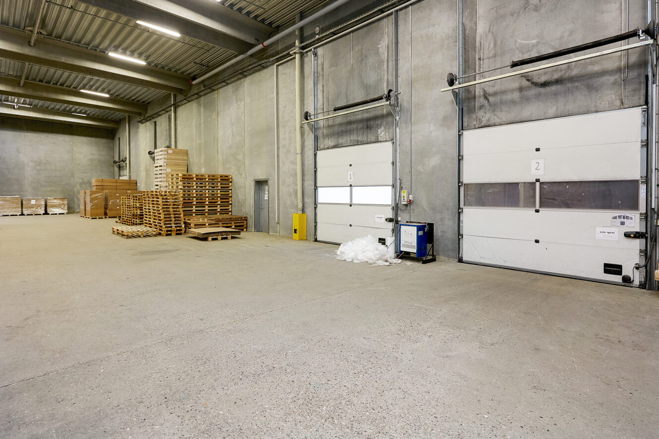 Interiør i en rummelig lagerhal med betonvægge, stablede træpaller, to store rulleporte og spredt hvid plastindpakning på gulvet.