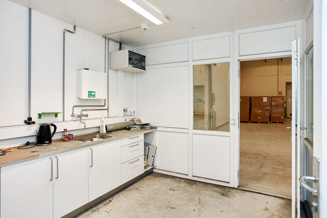 Et lille tekøkken med hvide skabe, vask, kedel og bordplade, der støder op til et industrielt rum med kasser, der ses gennem en åben dør.