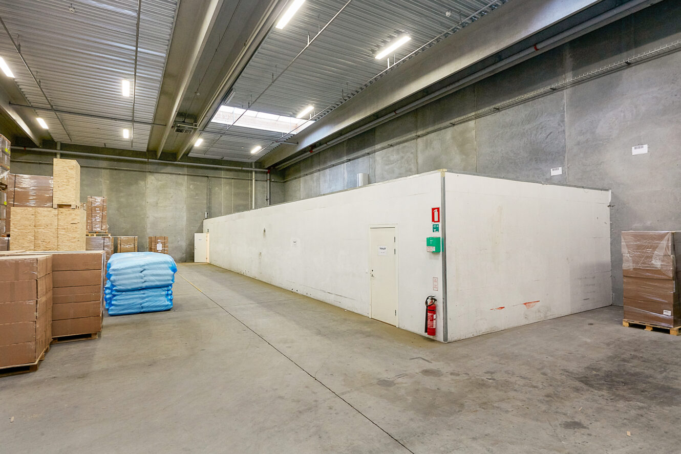 Lagerinteriør med højt til loftet, stablede paller, blå sække og et stort hvidt lukket rum i midten. Brandslukker og nødskilte er synlige på væggen.