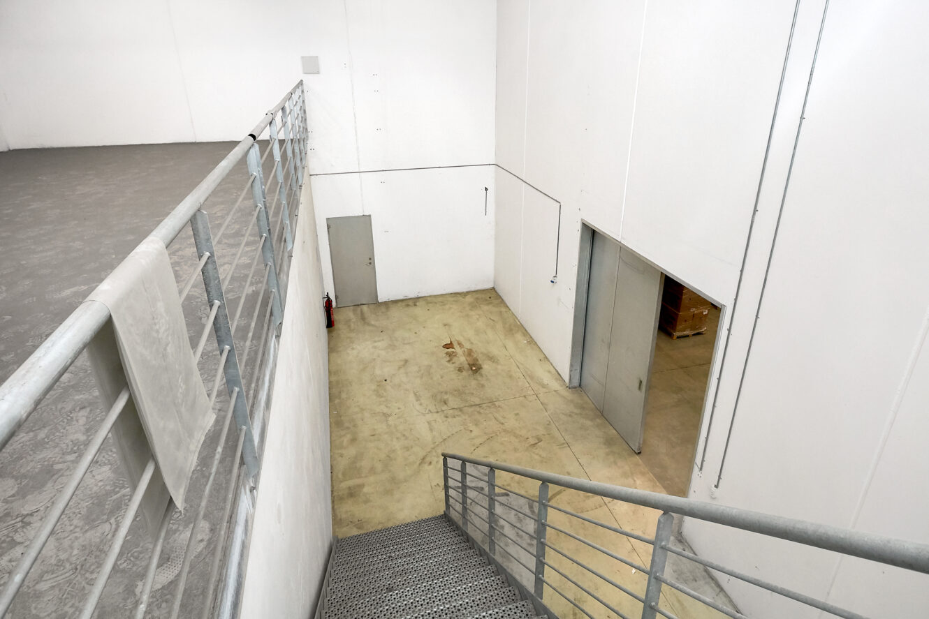 Et kig fra en metaltrappe, der viser et rent industrielt interiør med hvide vægge, betongulve, en brandslukker og en lukket dør i bunden.