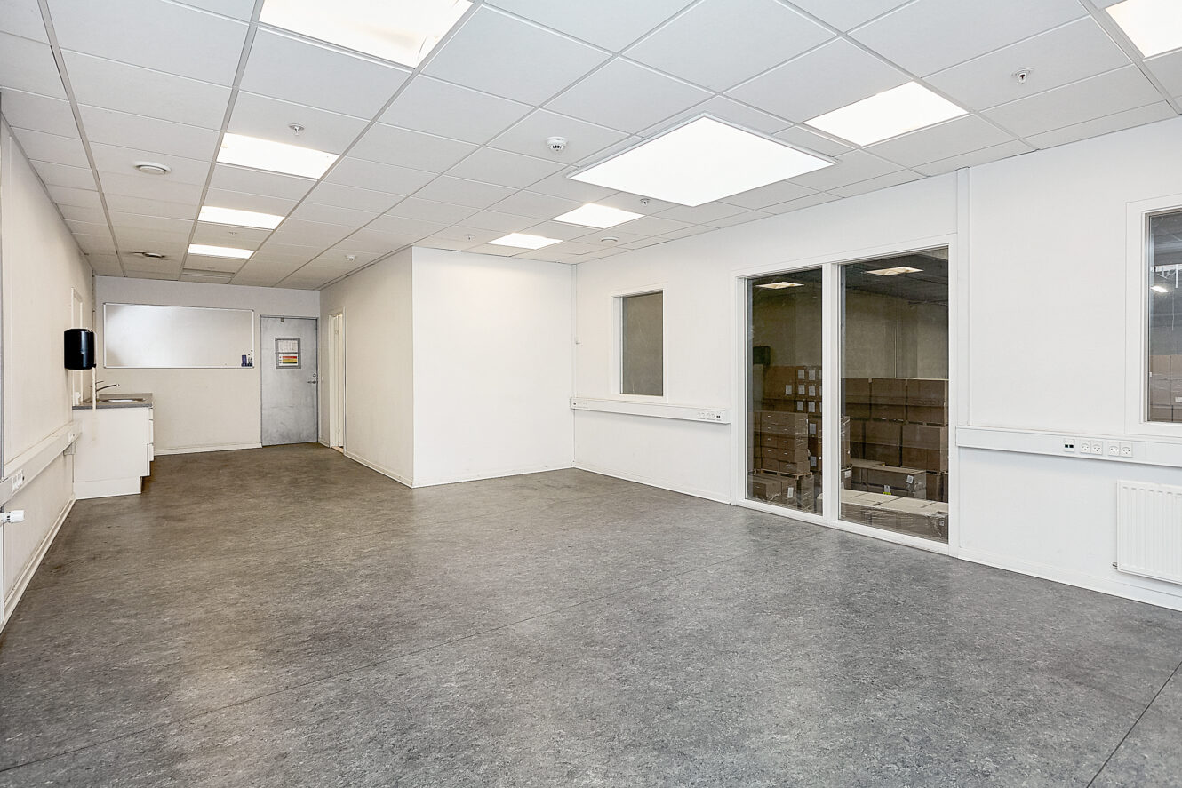 Et stort, tomt rum med hvide vægge, gråt gulv, loftslamper og vinduer med udsigt til et lagerområde med papkasser.