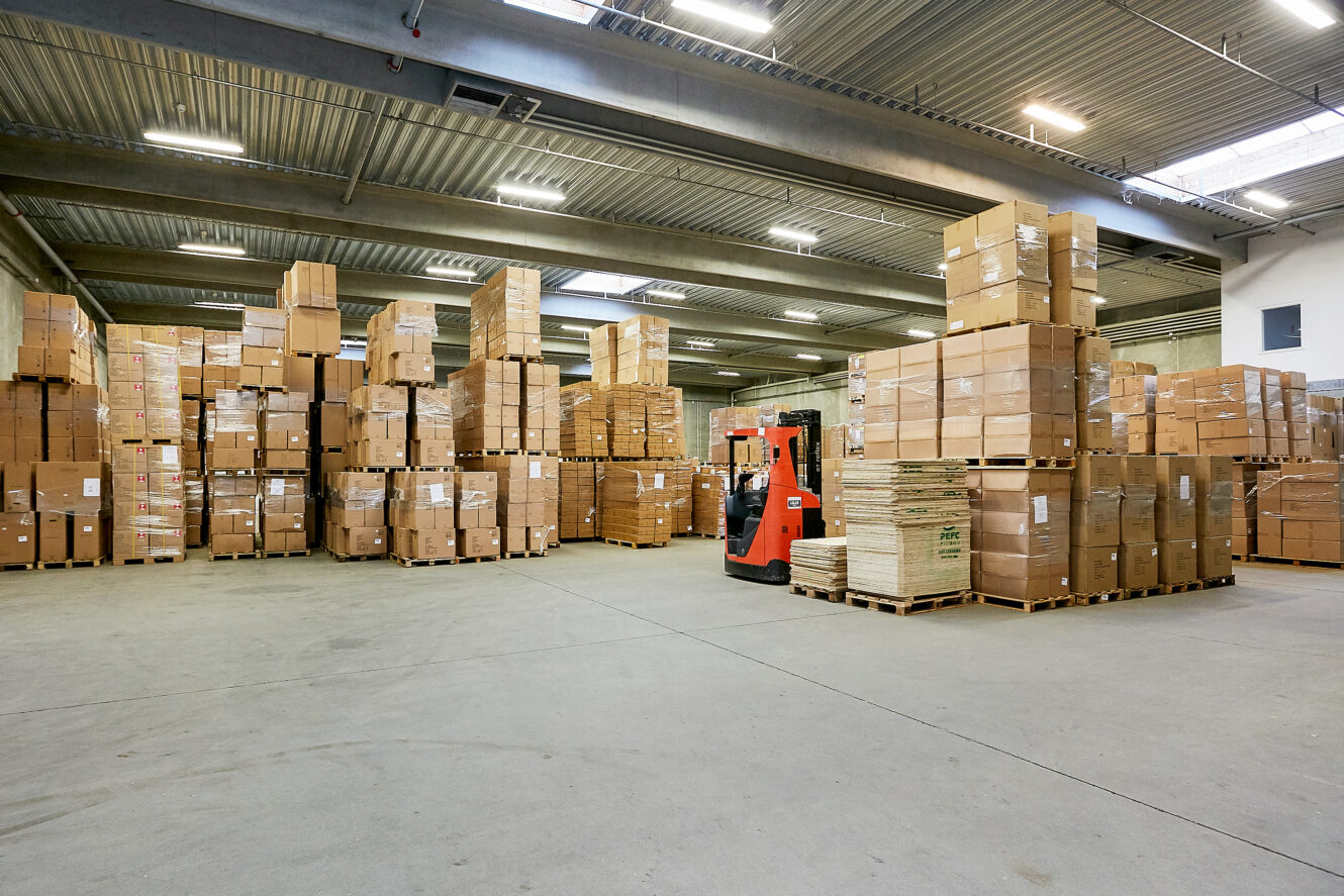 Stort lagerinteriør med stabler af papkasser på paller og en rød gaffeltruck, der kører i midten.