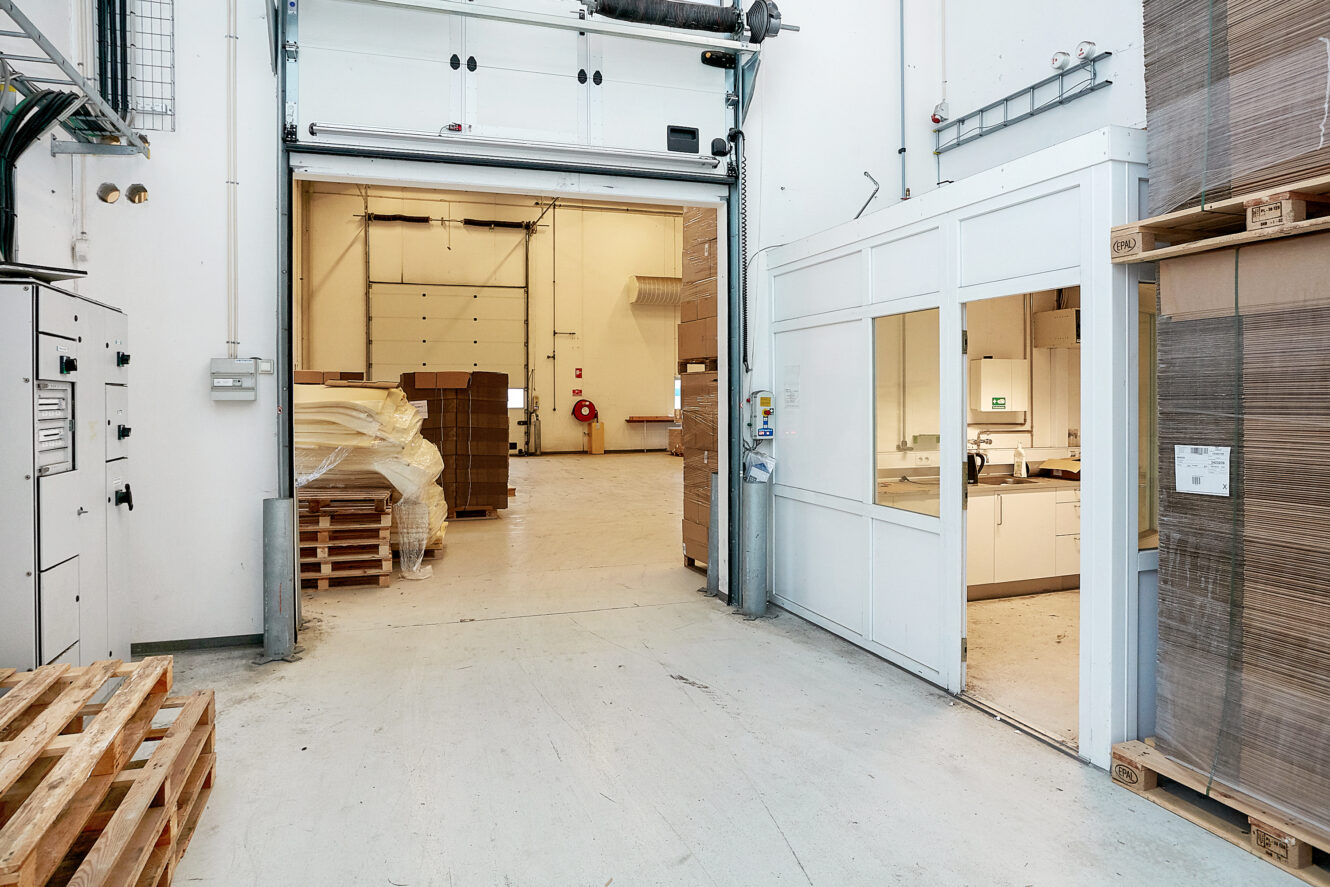 Et lagerinteriør med stablede paller, en delvist åben rulleport og et lille vindue, der afslører et kontor eller pauserum i højre side.
