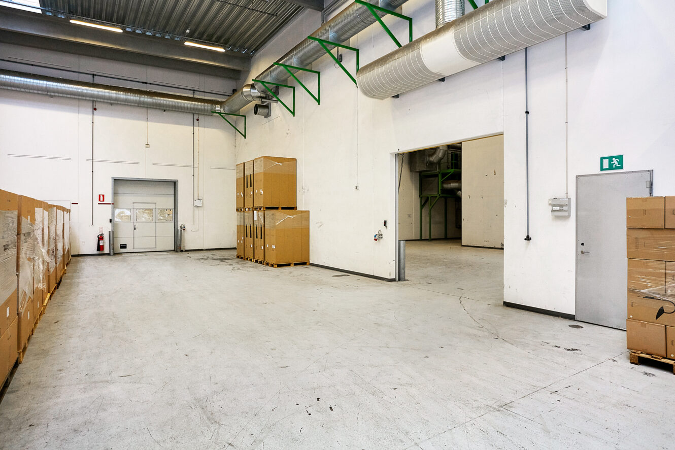 Et rent, rummeligt lagerinteriør med stabler af papkasser langs væggene, højt til loftet, synlige kanaler og en åben dør til et tilstødende rum.