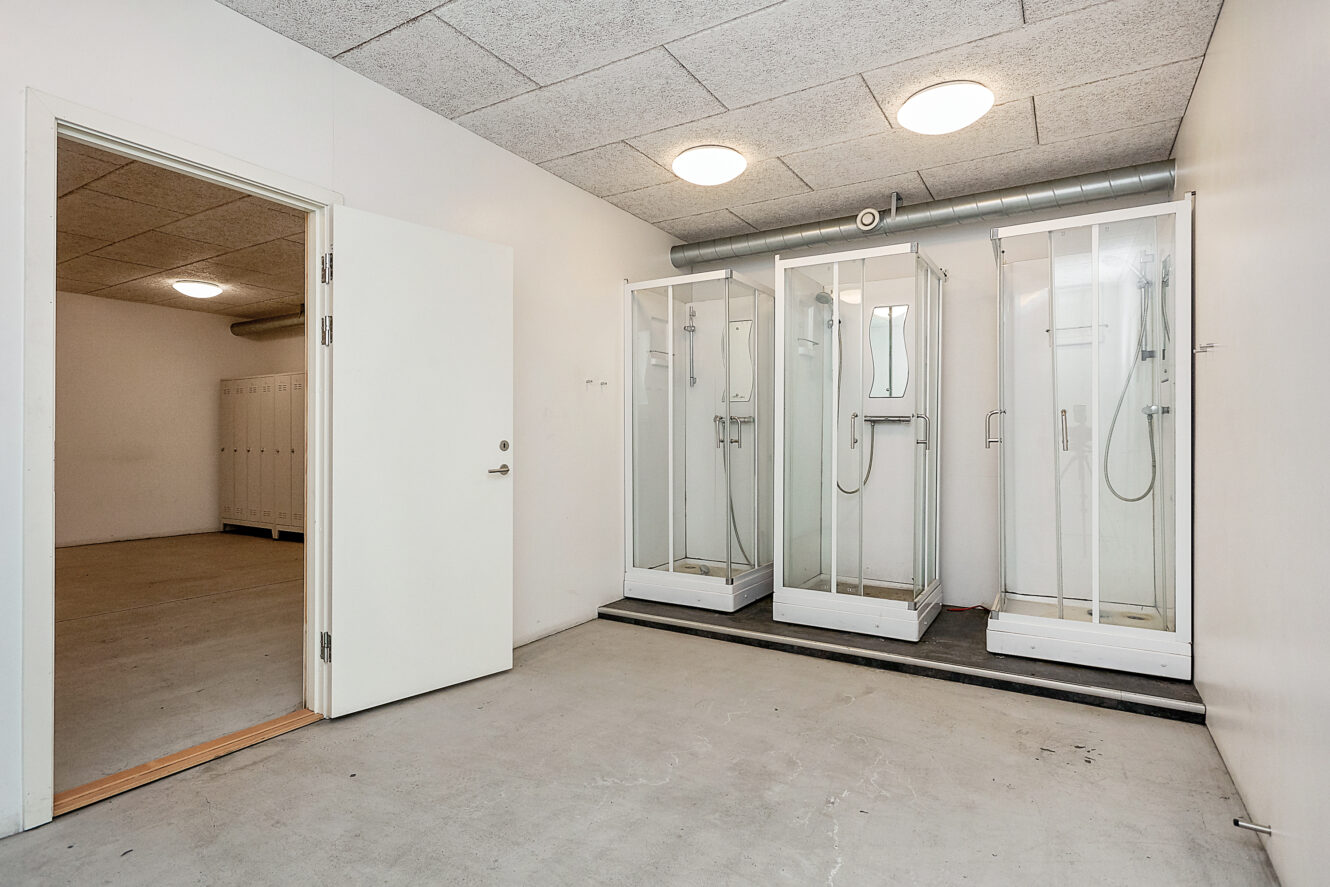 Et rum med tre brusekabiner af glas, en åben dør til et omklædningsområde, hvide vægge og et bart betongulv.