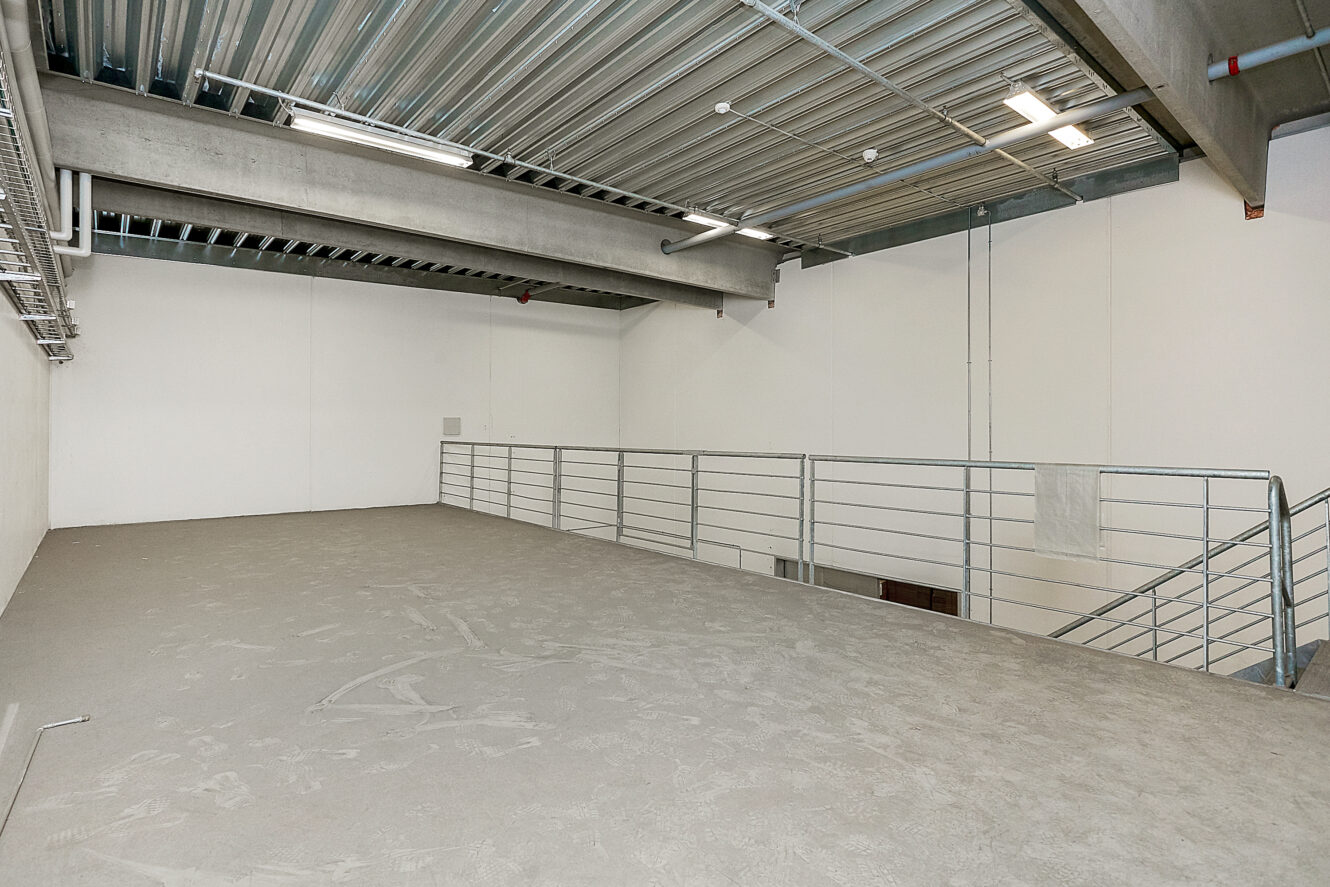Tom industriel mezzanin med betongulv, metalrækværk, synlige loftsbjælker og ovenlys i en lagerbygning eller et forretningslokale.