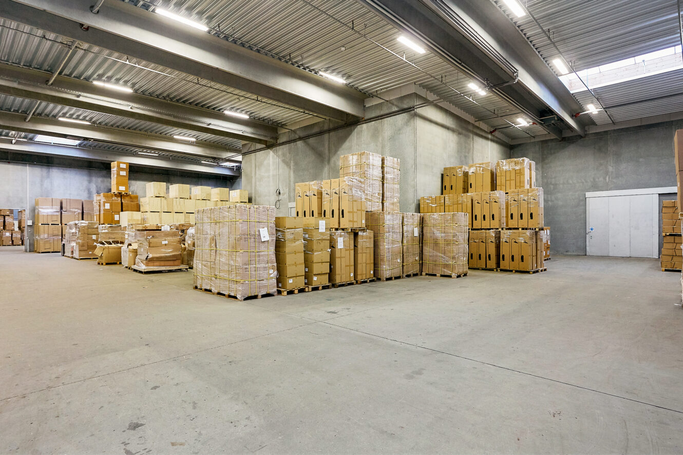 En stor lagerhal med betonvægge, højt til loftet og stabler af papkasser på træpaller langs væggene.