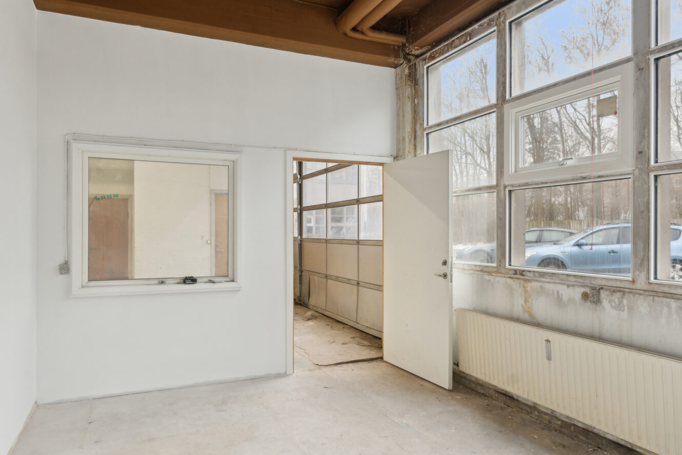 Tomt rum med et stort vindue, en åben dør, der fører til et andet område, en radiator og slidte vægge. En bil er synlig udenfor gennem vinduerne.