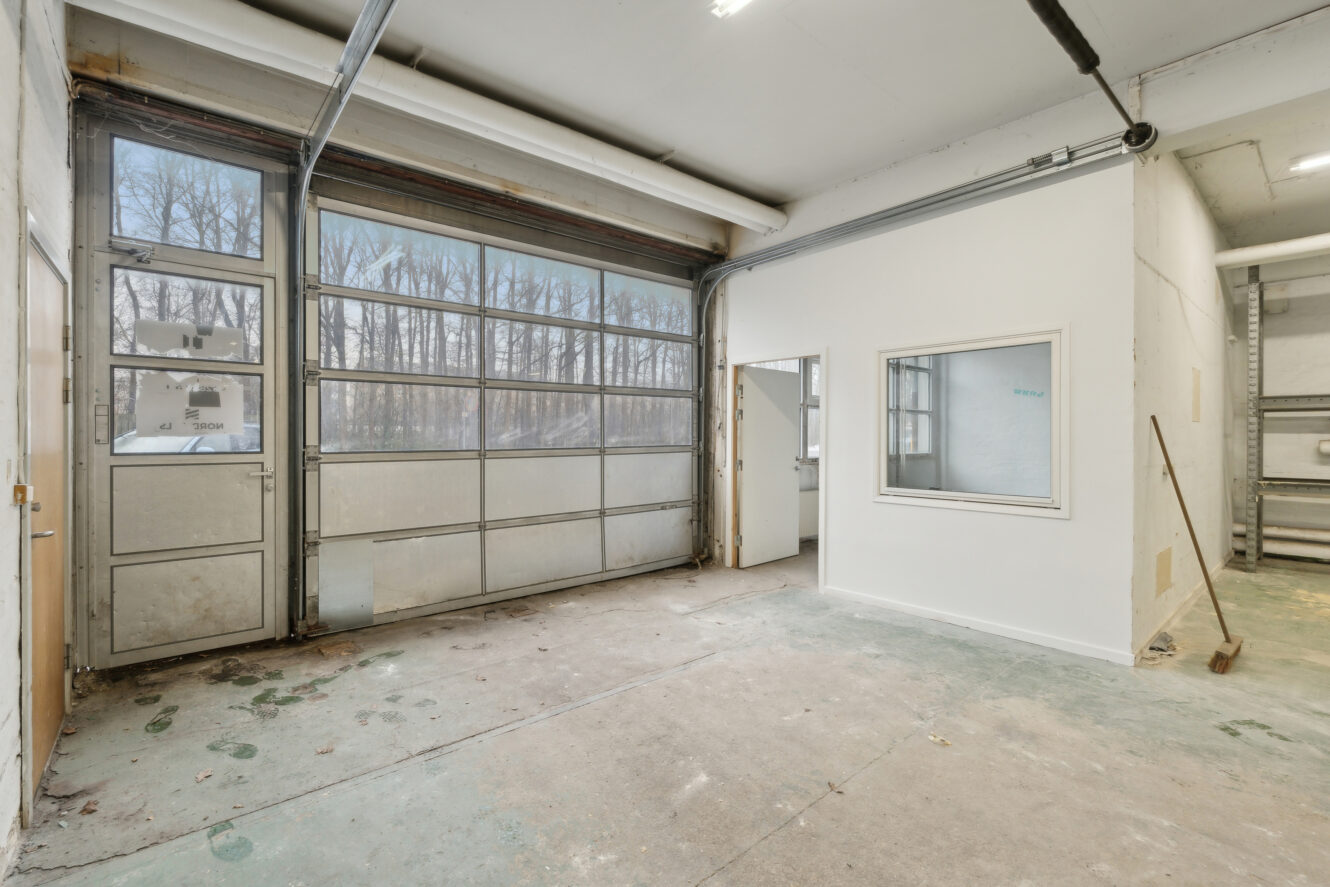 En tom industrigarage med en stor glasdør, et lille kontorlokale med et vindue og betongulve med snavs og fodaftryk.