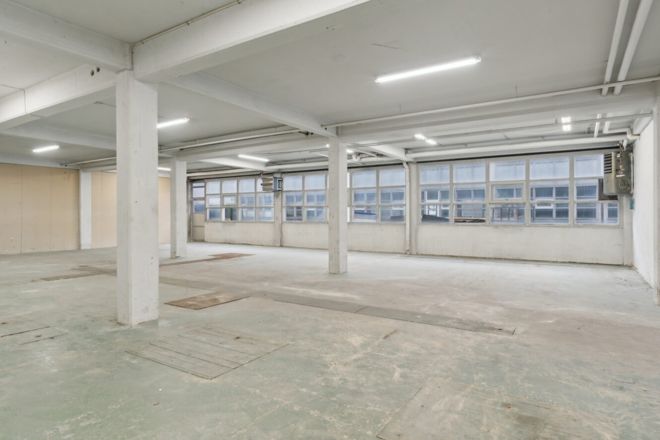 Rummeligt, tomt industrilokale med hvide søjler, store vinduer langs den ene væg, synlige rør og lysstofrør i loftet.